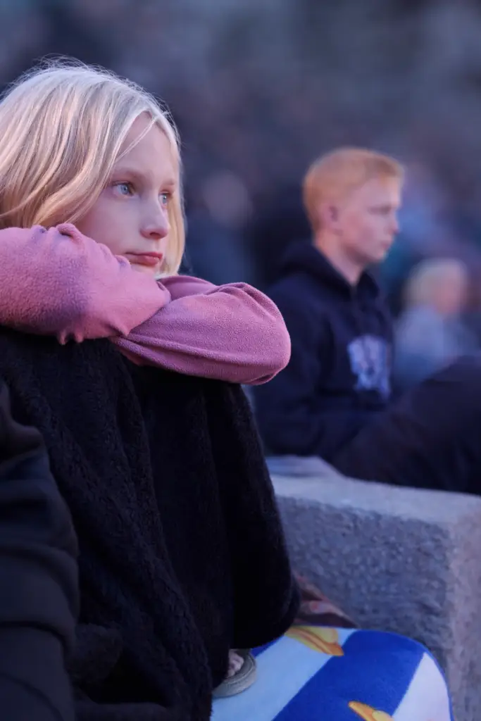 Ung blond pige sidder med armene omkring knæene i skumringen under teaterforestilling ved Minach Teatret.